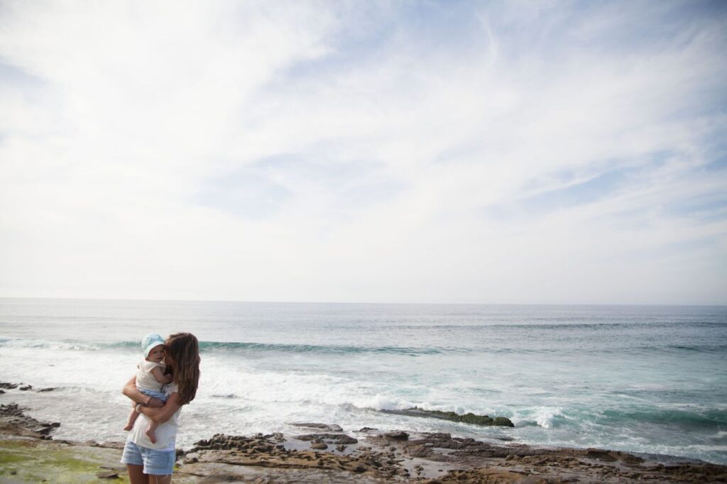 about-us-02 A mother embraces her child on a scenic beach with the ocean in the background.