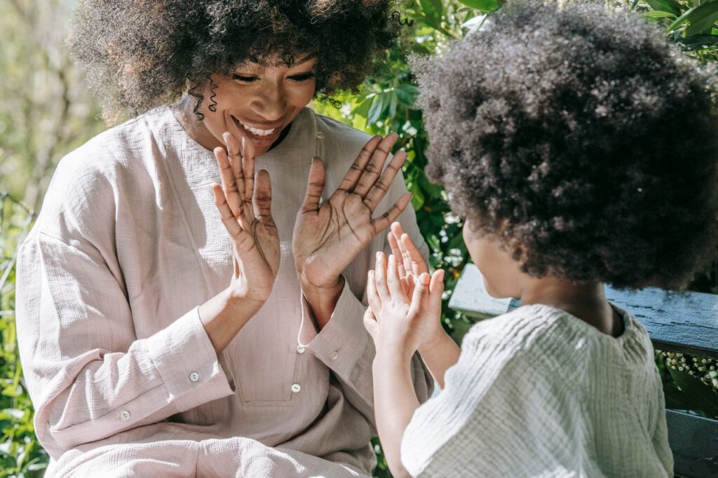 home-hero-1 Mother and daughter share a moment playing clapping game outdoors. Vibrant and joyful interaction.